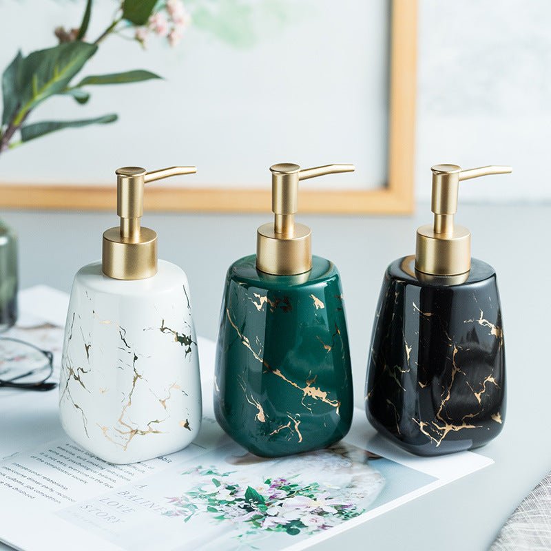 Three decorative pump bottles with a glossy marble finish are lined up on a tabletop. The bottles are white with gold veining, dark green with gold veining, and black with gold veining, all featuring gold-tone pump dispensers.
