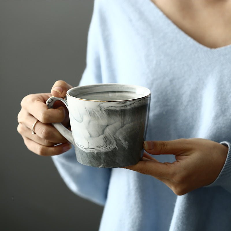 A person in a light blue sweater holds a ceramic mug with a gray and white marbled pattern and a thin gold rim against a dark gray background.