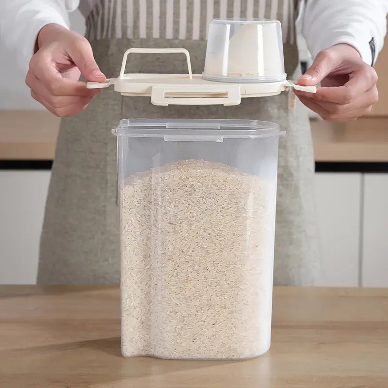 A person is holding a transparent sealed food storage container with a beige lid, filled with grains. The container is placed on a wooden table, and the person is in a kitchen setting.