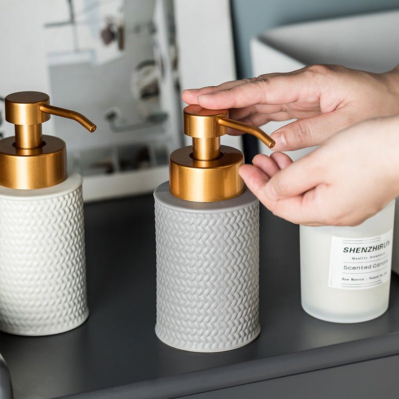 Person using a gold pump on a white soap dispenser with a blurred bathroom background