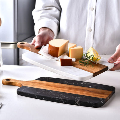 Person holding a wooden cutting board with cheese and herbs on a kitchen counter.