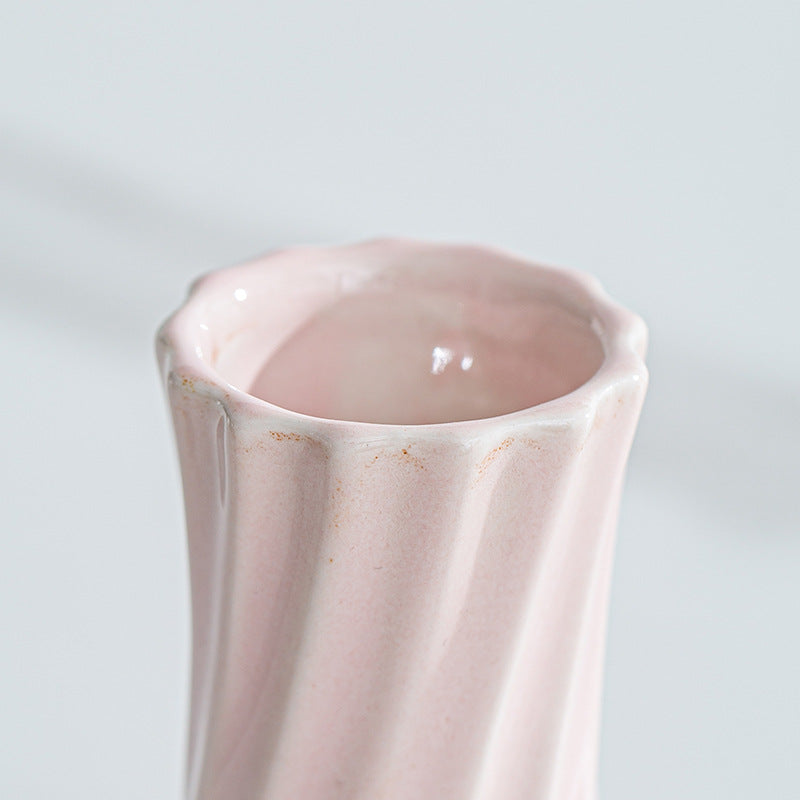 Close-up of a pale pink ceramic vase with a fluted, ribbed exterior and a scalloped rim, shown against a light gray background.