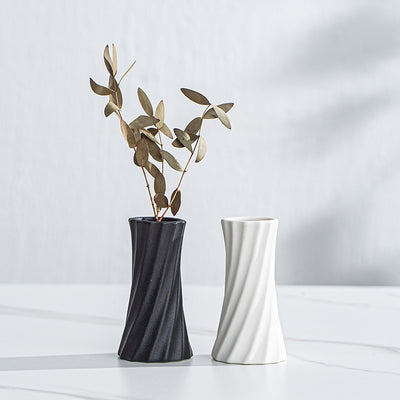 Two modern twisted ceramic vases on a white marble surface against a light gray wall. The left vase is dark charcoal gray and contains dried gold-toned eucalyptus stems, while the right vase is matte white.