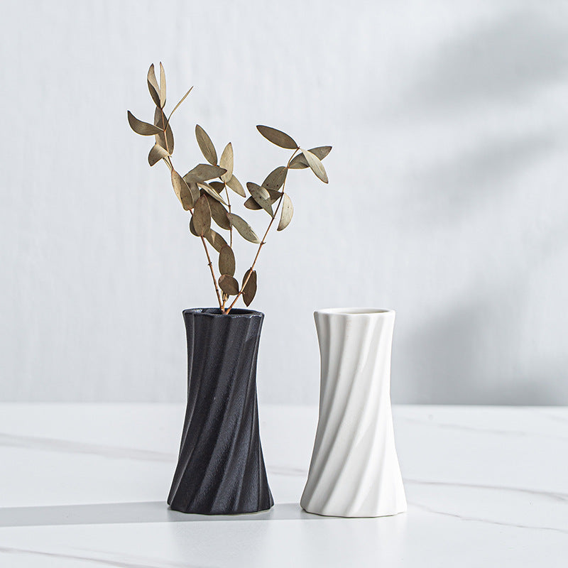 Two modern twisted ceramic vases on a white marble surface against a light gray wall. The left vase is dark charcoal gray and contains dried gold-toned eucalyptus stems, while the right vase is matte white.