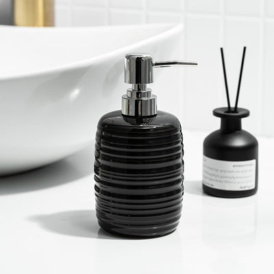 Black ribbed soap dispenser with silver pump on a white surface, next to a black diffuser bottle.