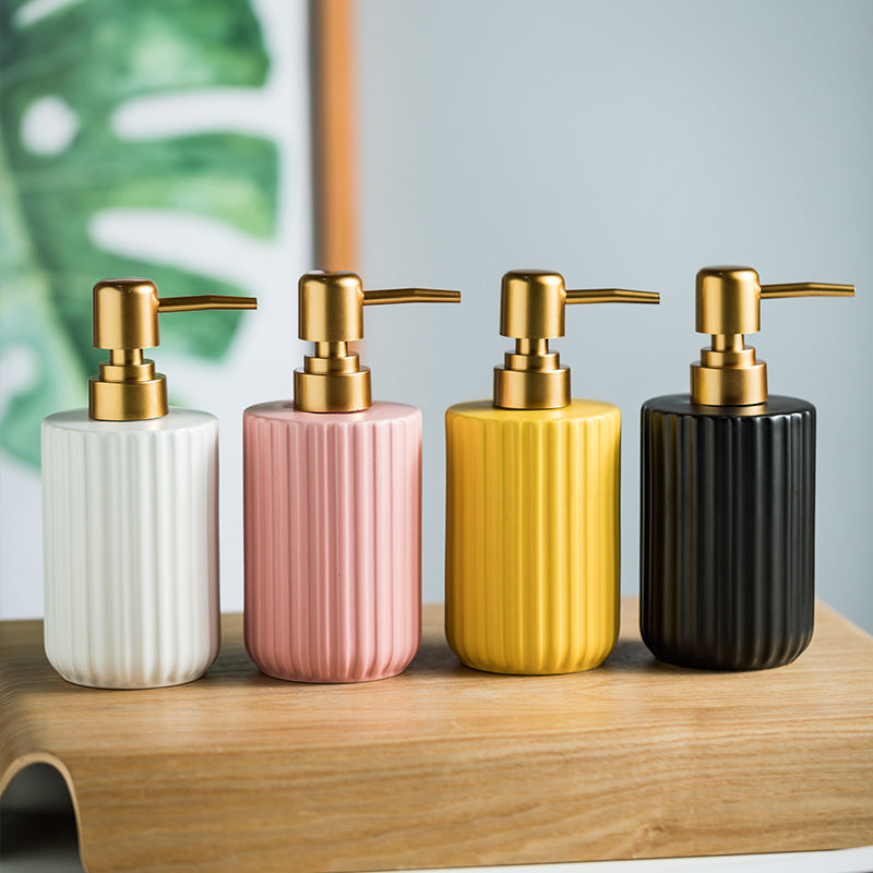 Four ceramic soap dispensers with gold pumps on a wooden surface, with a plant in the background.