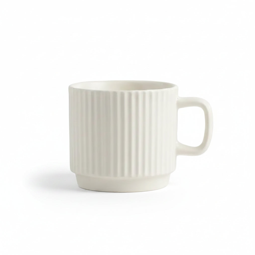 An off-white ceramic mug with a vertical ribbed texture and a C-shaped handle, set on a white textured fabric surface with a decorative cream-colored bubble candle in the background.
