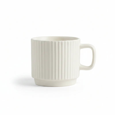 An off-white ceramic mug with a vertical ribbed texture and a C-shaped handle, set on a white textured fabric surface with a decorative cream-colored bubble candle in the background.