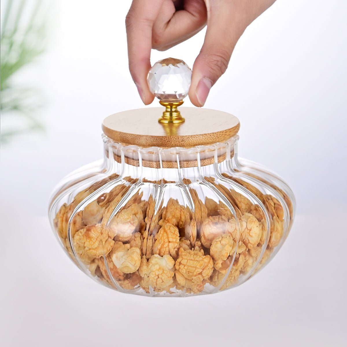 Glass jar with wooden lid filled with snacks, hand reaching to open it on a white background