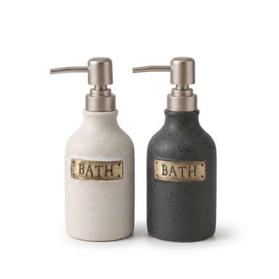 Two ceramic bath pump bottles on a white background, one in speckled cream with a brass plate reading BATH and one in speckled charcoal with a brass plate reading BATH.