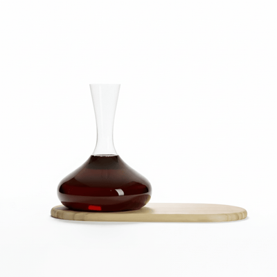 A clear glass decanter with a bulbous base, partially filled with dark red liquid, resting on a light natural wood oval board against a solid white background.