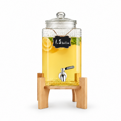 A 1.5 gallon glass beverage dispenser with a clear lid, filled with yellow lemonade and shown on a light wood stand against a warm cream background.
