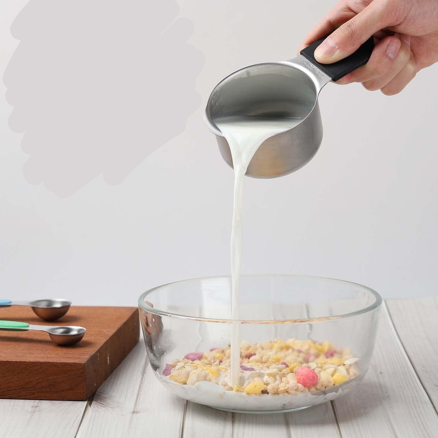Stainless steel measuring cup pouring milk into a bowl, with measuring spoons on a wooden board.