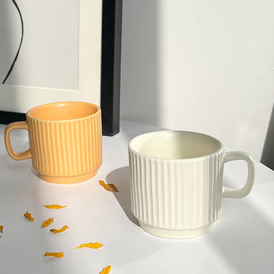 Two ribbed ceramic mugs on a white surface, one in warm yellow-orange and one in off-white, with a few scattered yellow petals in front. A black frame and white wall are visible in the background.
