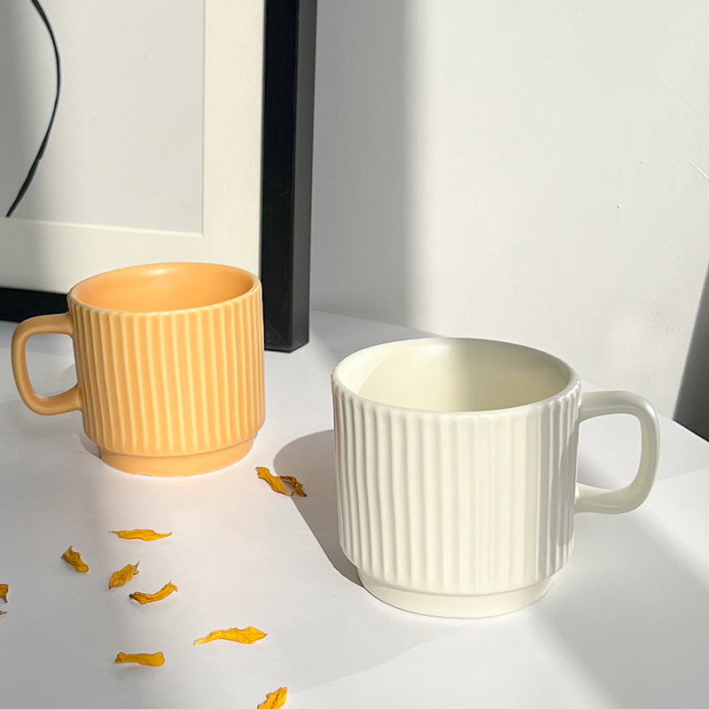 Two ribbed ceramic mugs on a white surface, one in warm yellow-orange and one in off-white, with a few scattered yellow petals in front. A black frame and white wall are visible in the background.